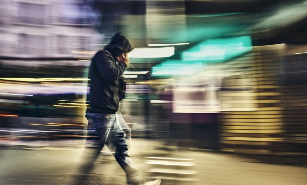 photographe Photo de style de vie nocturne prise par un photographe à Paris : une personne vêtue d’une veste sombre et d’un jean marche dans une rue de la ville éclairée par des lumières vives, avec les bâtiments emblématiques de Paris en arrière-plan ; cette image floue rend bien l’ambiance urbaine parisienne la nuit. Paris
