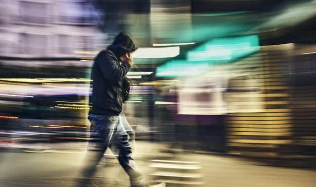 photographe Photo de style de vie nocturne prise par un photographe à Paris : une personne vêtue d’une veste sombre et d’un jean marche dans une rue de la ville éclairée par des lumières vives, avec les bâtiments emblématiques de Paris en arrière-plan ; cette image floue rend bien l’ambiance urbaine parisienne la nuit. Paris