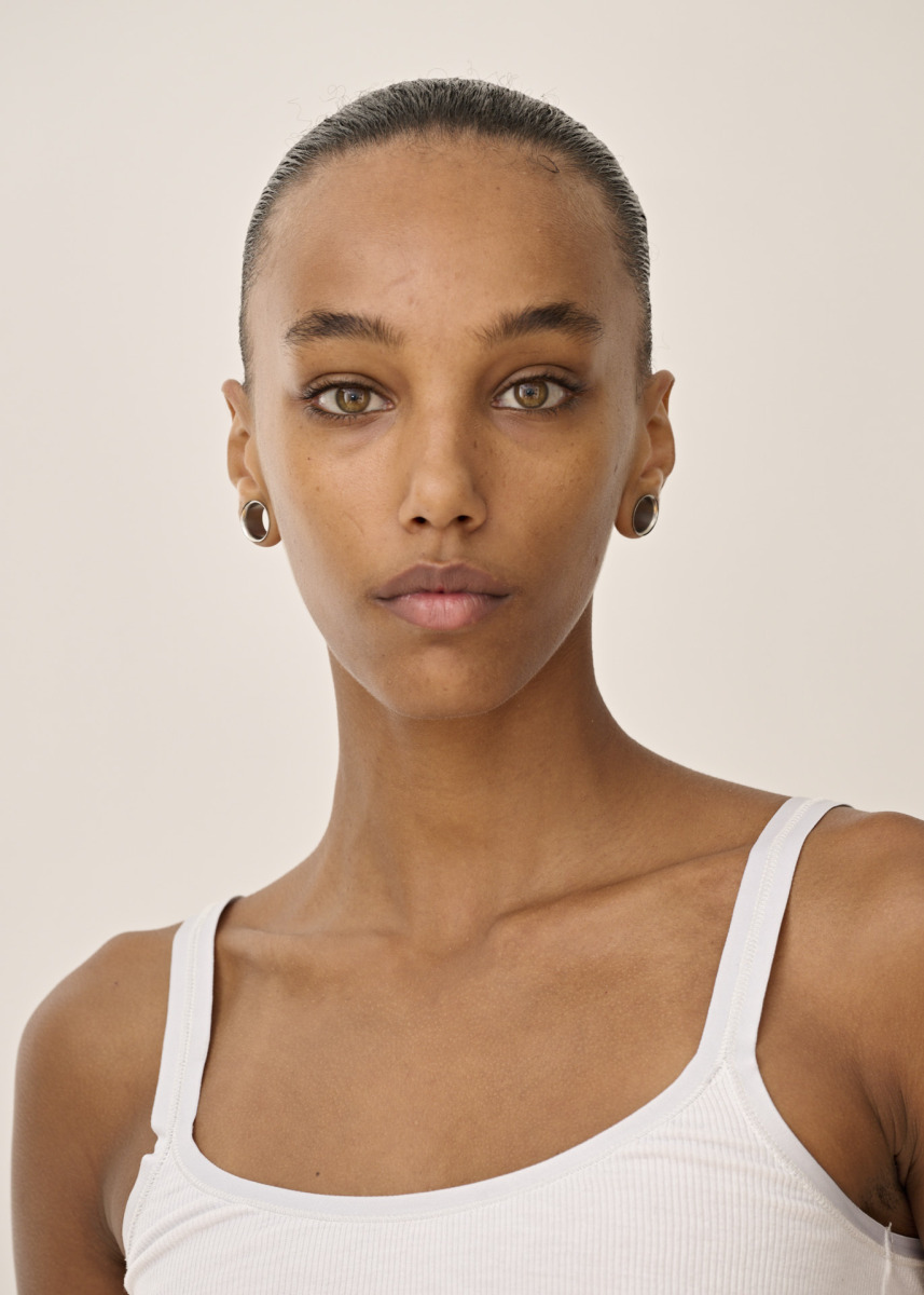photographe Portrait réalisé par un photographe à Paris : une personne à la peau beige clair, aux yeux noisette et aux cheveux lissés en arrière, porte un débardeur blanc et des boucles d’oreilles rondes. Elle regarde l’objectif avec une expression neutre, devant un fond uni, dans le cadre d’une séance photo de beauté ou pour un book de mannequin. Paris