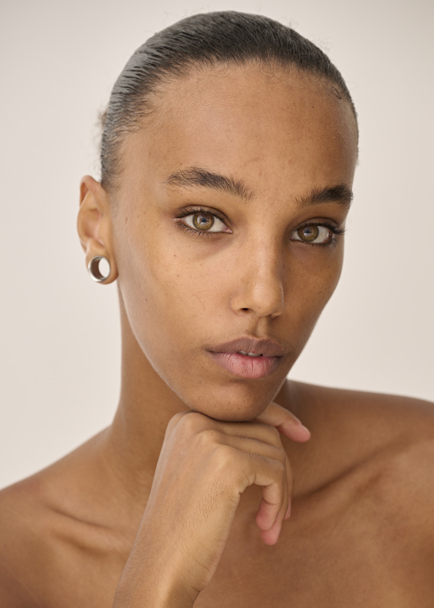 photographe Portrait de mode à Paris réalisé par un photographe professionnel : une personne à la peau claire, aux yeux noisette clairs et aux cheveux foncés courts tirés en arrière, regarde l’objectif avec une expression calme et neutre, le menton posé sur une main. Elle porte une petite boucle d’oreille discrète. Idéal pour un book de mannequin ou une séance photo lifestyle à Paris. Paris