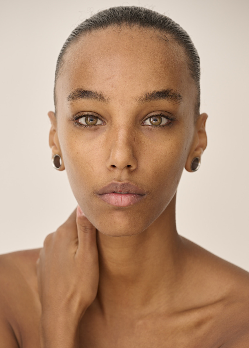 photographe Portrait d’une photographe à Paris : une personne à la peau mate, aux yeux noisette et aux cheveux foncés tirés en arrière, posant face à l’objectif avec une expression neutre et une main posée sur son cou. Elle porte des plugs noirs aux deux oreilles. L’arrière-plan est clair et épuré, idéal pour un book de mannequin ou une photo de beauté à Paris. Paris