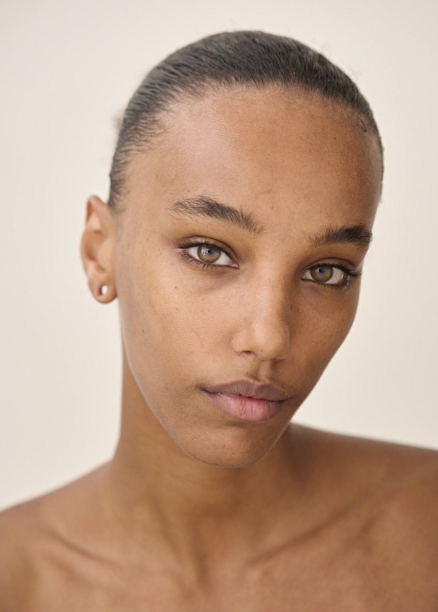 photographe Portrait de beauté réalisé par un photographe professionnel à Paris, mettant en valeur une personne aux yeux marron clair et à la peau lisse, regardant l’objectif avec une expression neutre. Les cheveux sont tirés en arrière et une petite boucle d’oreille clou est visible. Le fond est uni et de couleur claire, idéal pour un book de mannequin ou des photos de mode en studio à Paris. Paris