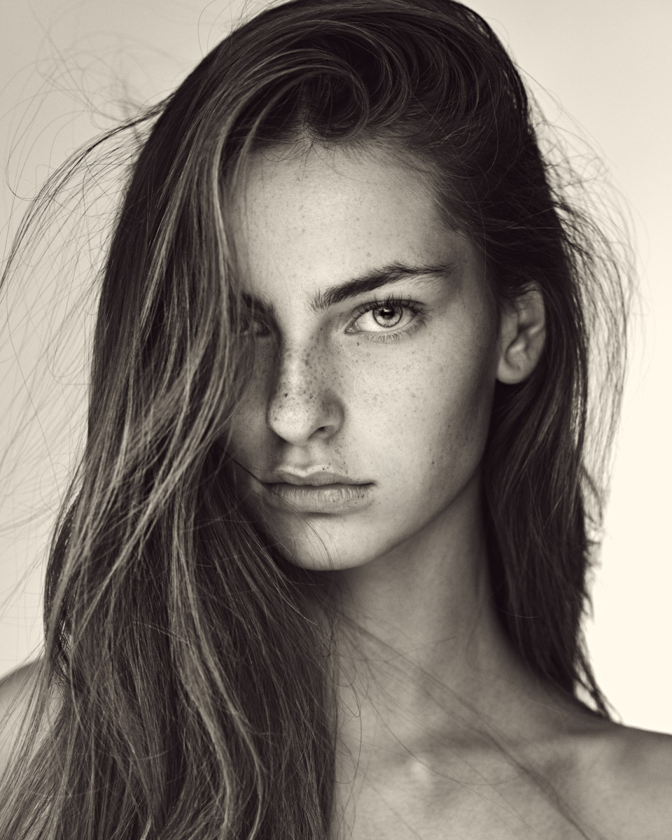 photographe Portrait en noir et blanc réalisé par un photographe à Paris : une jeune personne aux cheveux longs et aux taches de rousseur pose avec sérieux devant l’objectif. Cette photo de mode met en valeur la lumière naturelle sur un fond uni, ce qui en fait un choix idéal pour un book de mannequin ou des photos lifestyle professionnelles. Paris