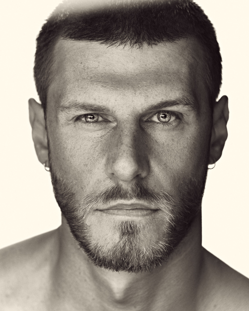 photographe Portrait en noir et blanc d’un photographe à Paris, pris en gros plan ; il a les cheveux courts, une barbe soignée, les yeux clairs et de petites boucles d’oreilles. Il regarde l’objectif avec une expression neutre, dans un style de portrait professionnel. Idéal pour un book de mannequin, de mode ou de lifestyle à Paris. Paris